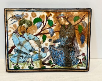 May include: Rectangular ceramic tile with a detailed illustration of two figures in traditional attire, seated beneath a tree with green leaves and blue fruit. The figures are in shades of blue, brown, and green, with a brown and tan background.