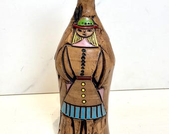 May include: A ceramic vase with a brown glaze and a painted design of a woman wearing a green hat, pink shirt, and blue skirt.