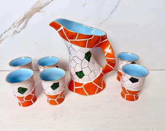 May include: A ceramic pitcher and six matching glasses. The set features a mosaic-like design with orange, white, and green accents. The interiors of the pitcher and glasses are light blue, with gold rims. A decorative drinkware set.