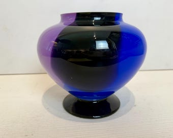 May include: A small, round glass vase with a purple, black, and blue color scheme. The vase has a wide base and a narrow neck.