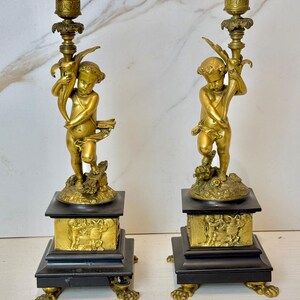 Puede incluir: Un par de candelabros antiguos de latón con figuras de querubines que sostienen una rama frondosa. Cada candelabro tiene una base cuadrada negra con detalles dorados y se apoya en cuatro patas de garra.