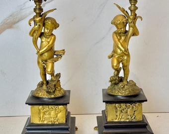 May include: A pair of antique brass candlesticks featuring cherub figures holding a leafy branch. Each candlestick has a black square base with gold accents and sits on four paw feet.