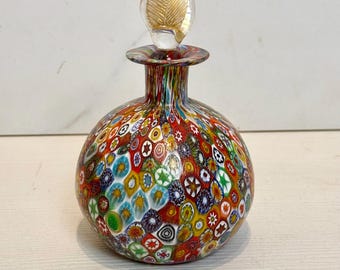 May include: A small, round glass bottle with a colorful, multi-patterned design. The bottle has a clear glass stopper with a gold leaf design.