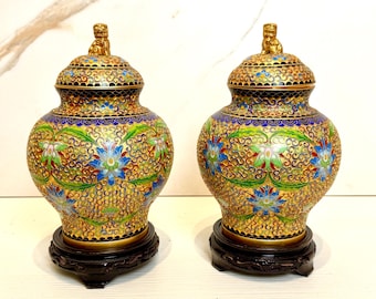 May include: A pair of decorative cloisonné vases with intricate floral patterns in shades of blue, green, and gold. The vases have a gold finish and are topped with lion finials. They are displayed on wooden stands.