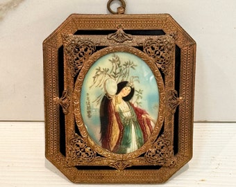 May include: A small, framed painting of a woman in a traditional Chinese dress holding a round mirror. The frame is made of gold metal and has a black background. The painting is surrounded by a decorative, ornate border.