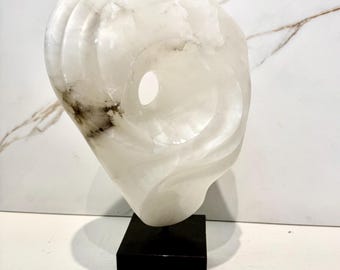 May include: White alabaster sculpture with a hole in the center, mounted on a black base.