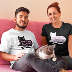 May include: White and black t-shirts with a cat graphic and the text "True love is fur-ever". A gray and white cat is sitting on a person's lap. The people are sitting on a pink couch.