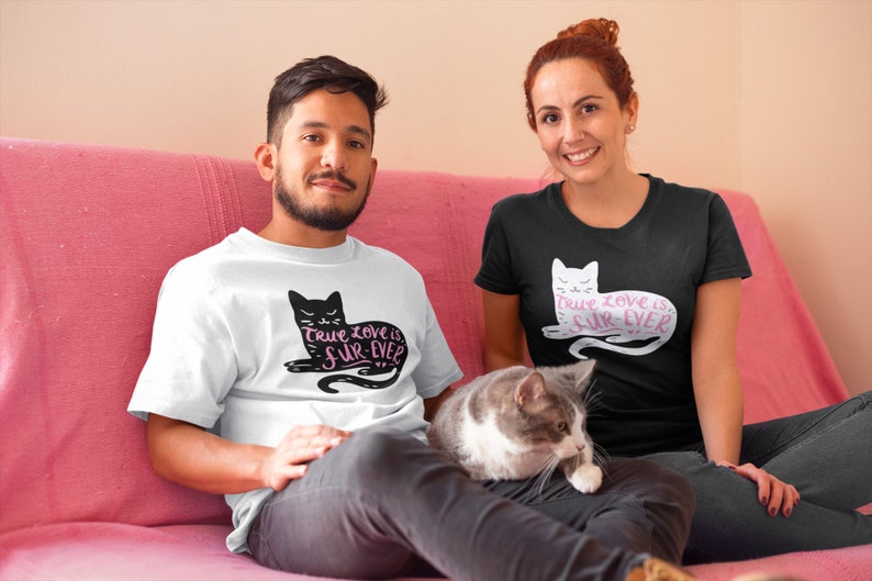 May include: White and black t-shirts with a cat graphic and the text "True love is fur-ever". A gray and white cat is sitting on a person's lap. The people are sitting on a pink couch.
