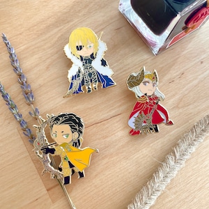 May include: Three enamel pins with anime-style character designs. One pin shows a blonde character with a blue cape and eye patch. Another features a character in a red cape. The third pin has a character with a bow and arrow.