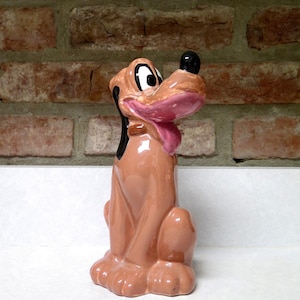May include: A ceramic figurine of Pluto, a cartoon dog. The figurine is primarily tan with a black nose, ears, and a pink tongue. It is sitting upright with its tongue sticking out. The background is a brick wall.