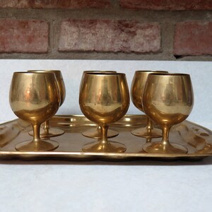 May include: Set of six gold-toned brass shot glasses on a matching brass tray. The glasses have a rounded, goblet-like shape.