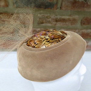 May include: A brown velvet hat with a brown feather trim. The hat has a veil attached to the back.