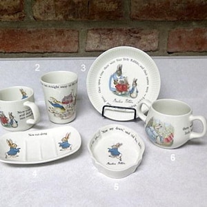 May include: A white ceramic dish set featuring illustrations of rabbits in blue jackets. The set includes a mug, a tumbler, a plate, a soap dish, and a small dish. The illustrations are from a children's book.