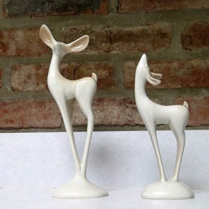 May include: Two off-white ceramic deer figurines standing on circular bases. The taller deer on the left has its head tilted upward with tan-colored inner ears, while the shorter deer on the right faces forward with a smooth, glossy finish.
