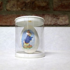 May include: An egg-shaped ornament featuring a rabbit in a blue jacket, set inside a clear plastic display case with a white base and top. The background includes a brick wall and a white surface.