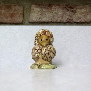 May include: A ceramic figurine of a mouse wearing a cloak. The mouse is light brown with black eyes and a red berry on its head. The cloak is beige and pink, and the base is green.