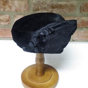 May include: A black hat with a rounded crown and a pleated brim. The hat features decorative fabric embellishments on the side. It is displayed on a wooden hat stand, showcasing its shape and design.
