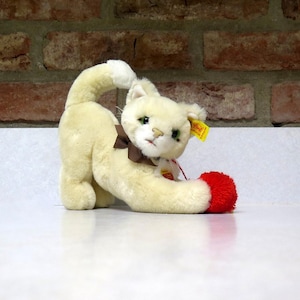May include: A cream-colored plush cat toy with a brown bow and a red ball. The cat is in a playful pose, with its tail arched and its paw touching the ball. The toy has green eyes and a yellow tag.