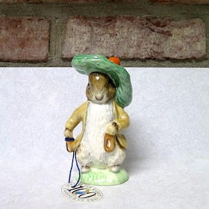 May include: A ceramic figurine of a rabbit wearing a green hat and a yellow coat. The rabbit is holding a small bag and has a tag attached. The figurine is standing on a patch of green grass.
