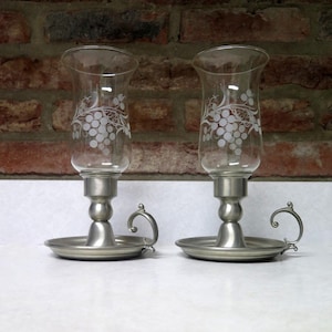 May include: Two clear glass candle holders with a silver-colored base and handle. The glass has a white grape vine design. The candle holders sit on a round base. The background is a brick wall.
