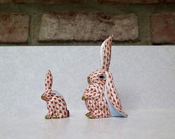 May include: Two porcelain rabbit figurines, one larger than the other. Both are white with a brown and white geometric pattern. The larger rabbit has its ears up and the smaller rabbit has its ears down.