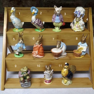 May include: A collection of eleven ceramic figurines of storybook characters, including rabbits, geese, a pig, and a frog, displayed on a tiered wooden shelf. The figurines are painted in various colors and dressed in detailed clothing, with each character posed differently.