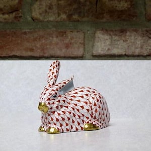 May include: A small, red and white porcelain rabbit figurine with gold accents. The rabbit is sitting on a white surface.