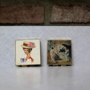 May include: Two vintage square compacts. One features a woman in a hat with pink roses, the other a painting of a ballerina. Both have gold-toned metal frames and are displayed on a white surface.