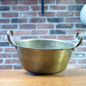 May include: A large, round, polished brass basin featuring two curved metal handles attached to opposite sides. The bowl has a smooth, slightly weathered surface with visible variations in the metallic finish and a wide, open top rim.