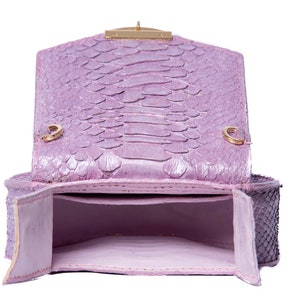 Pu&ograve; includere: Una borsa a mano in pelle di serpente color lavanda con una chiusura dorata e una patta che si apre per rivelare un interno rosa.