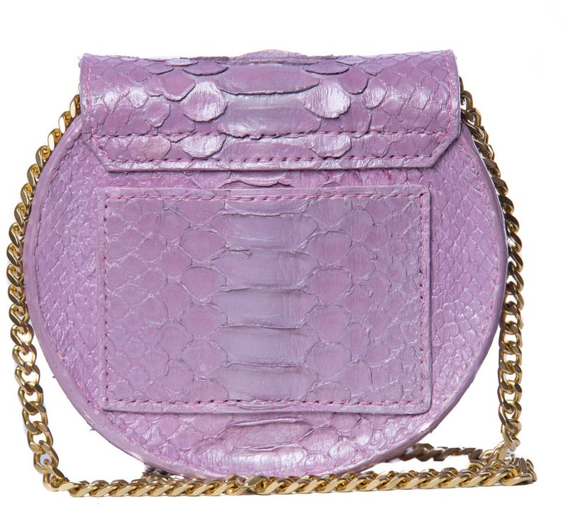 Pu&ograve; includere: Una piccola borsa rotonda di colore lavanda con una tracolla a catena dorata. La borsa &egrave; realizzata in pelle di serpente e ha una chiusura a lembo.