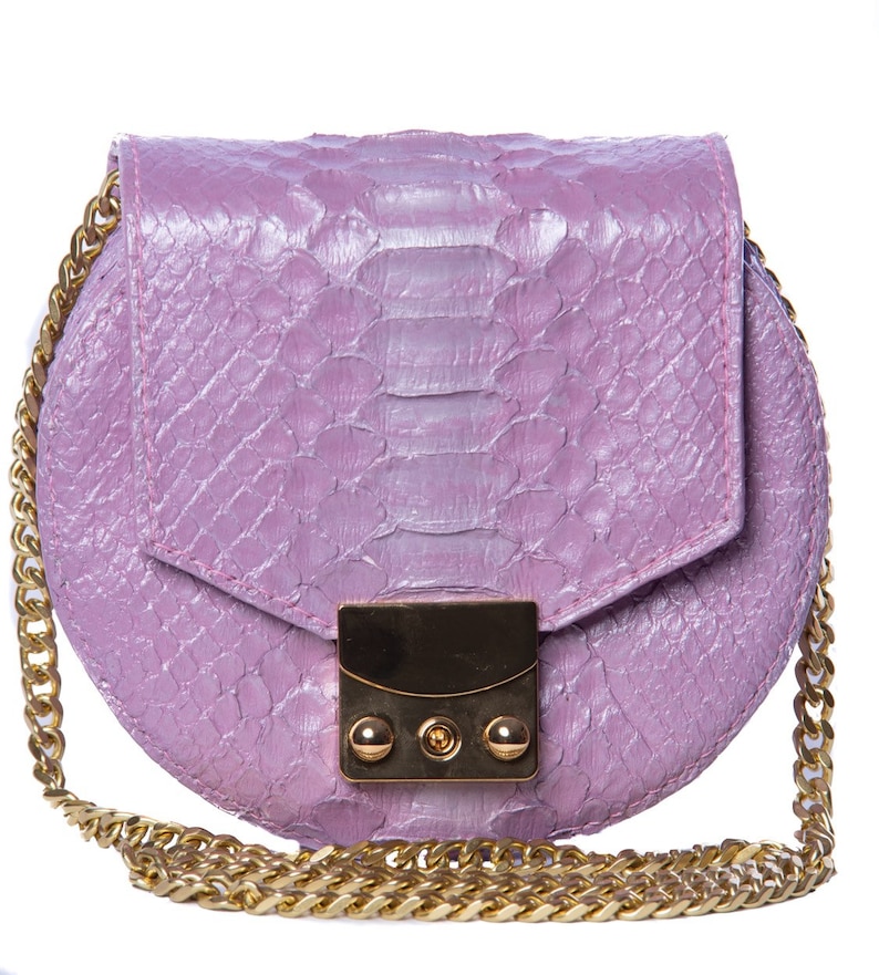 Pu&ograve; includere: Una piccola borsa rotonda di colore lilla chiaro con una tracolla a catena dorata e una chiusura dorata. La borsa &egrave; realizzata in pelle di serpente.