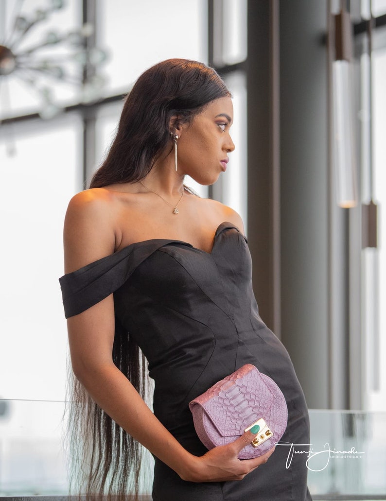 Pu&ograve; includere: Una donna con un abito nero scollato e che tiene una piccola pochette di pelle di serpente di colore lilla chiaro.