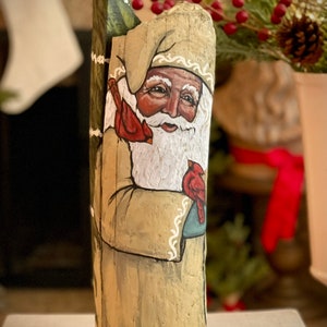 May include: A hand-painted wooden figurine of Santa Claus holding a cardinal bird. The figurine is painted in shades of brown, white, red, and green. The figurine is standing on a wooden surface.