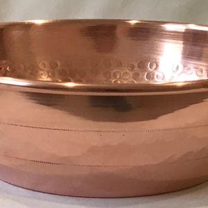 May include: A copper bowl with hammered texture and brass handles. The bowl has a rounded shape and a slightly flared rim.