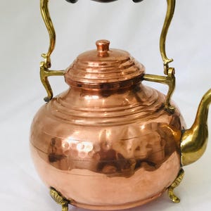 May include: A copper teapot with a hammered finish, a wooden handle, and brass accents. The teapot has a round body and a lid with a knob. It sits on three brass feet.