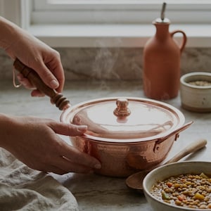 May include: Copper cooking pot with lid, wooden handle, and a small ceramic bowl. The image includes the text "Your Kitchen Essential". A linen cloth and a bowl of soup are also visible.