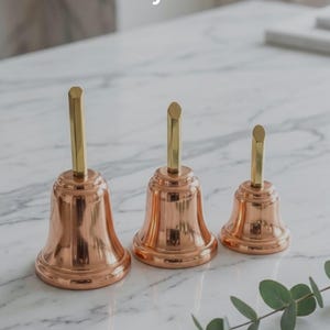 May include: Three copper bells with gold-colored handles are displayed on a marble surface. The bells vary in size, with the largest on the left and the smallest on the right. The text "Your Identity In Every Note" is at the top.