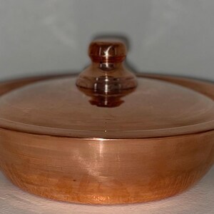 May include: A small copper pot with a lid and two handles. The pot is shiny and has a hammered finish.