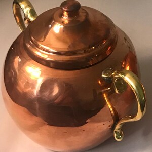 May include: A copper sugar bowl with a lid and two brass handles. The bowl is round and has a shiny finish.