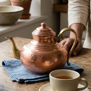 May include: A copper teapot with a brass handle and spout sits on a blue napkin. A teacup and saucer with a gold spoon are on a wooden table. A plant in a terracotta pot is in the background, suggesting a cozy kitchen setting.