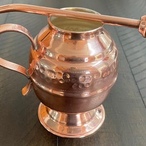 Könnte beinhalten: Eine Kupferkanne mit gehämmerter Oberfläche und einem Ausguss. Die Kanne hat einen Henkel und einen runden Boden. Sie wird mit einem Kupferstrohhalm mit einem kleinen Loch am Ende geliefert.