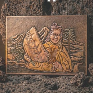 May include: A copper relief depicting a figure holding a drum, with mountains and trees in the background. The text reads "A thousand years on copper" and "The ancient spirit of the Araucaria tree, forged into an heirloom that outlasts time."
