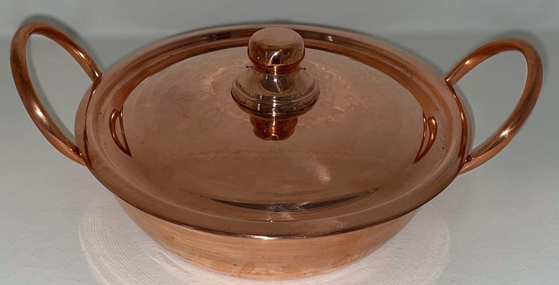 May include: A copper serving dish with a lid and two handles. The dish is round and has a shiny finish.