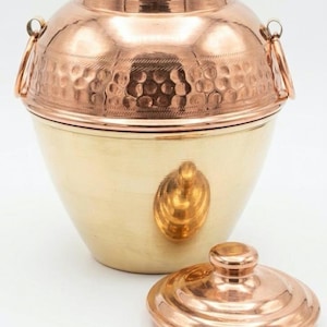 May include: A two-tone copper and brass container with a lid. The upper portion is copper with a hammered texture and two handles. The lower portion is brass with a decorative brass accent. The lid is copper.
