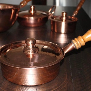 May include: A set of four copper pots with wooden handles. The pots have lids and are arranged on a dark wooden surface.