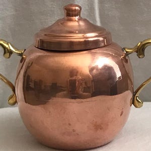 May include: A copper sugar bowl with a lid and two gold-colored handles. The bowl has a rounded shape and a shiny, reflective surface. The lid has a small knob on top. The handles are curved and ornate.