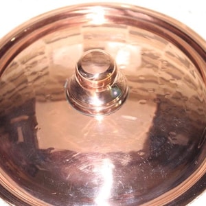 May include: A copper pot lid with a round knob on top. The lid is shiny and has a hammered finish.