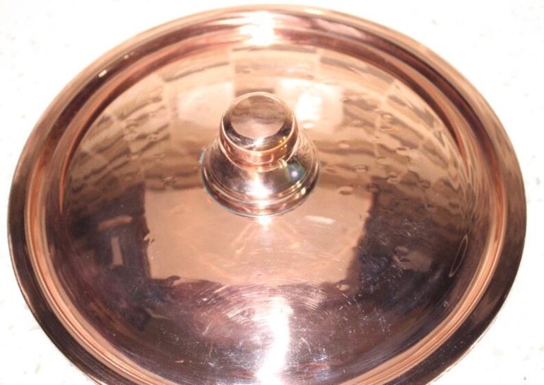 May include: A copper pot lid with a round knob on top. The lid is shiny and has a hammered finish.