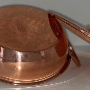 May include: A copper pot with a lid and two handles. The pot is shiny and has a rounded bottom.